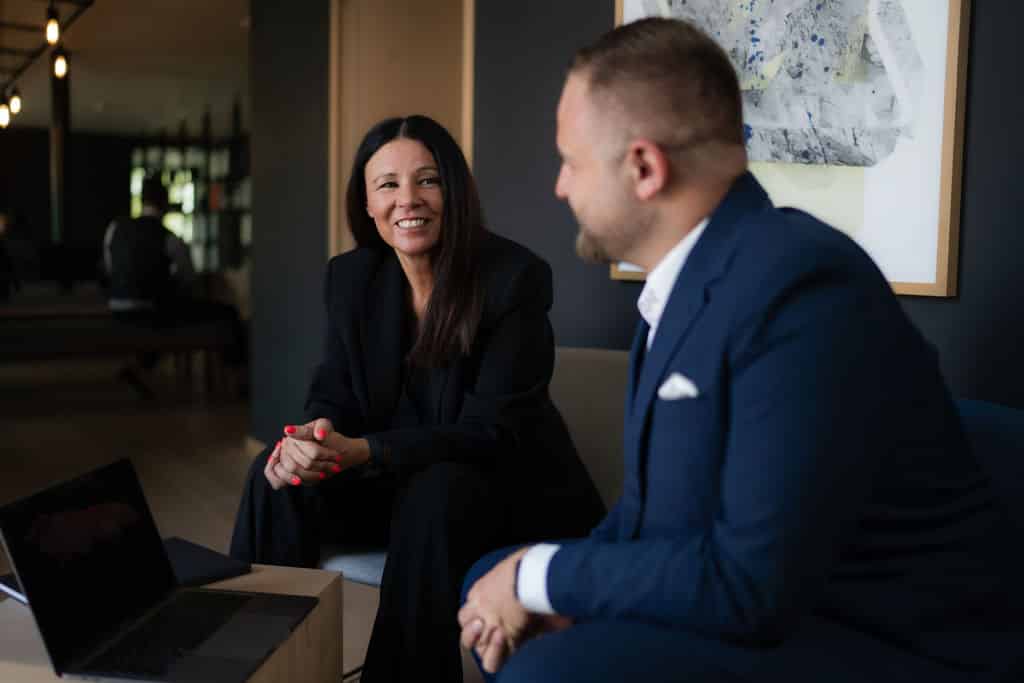 Troi Businessfoto: Zwei Personen im Gespräch in moderner Büro-Umgebung mit aufgeklapptem Laptop